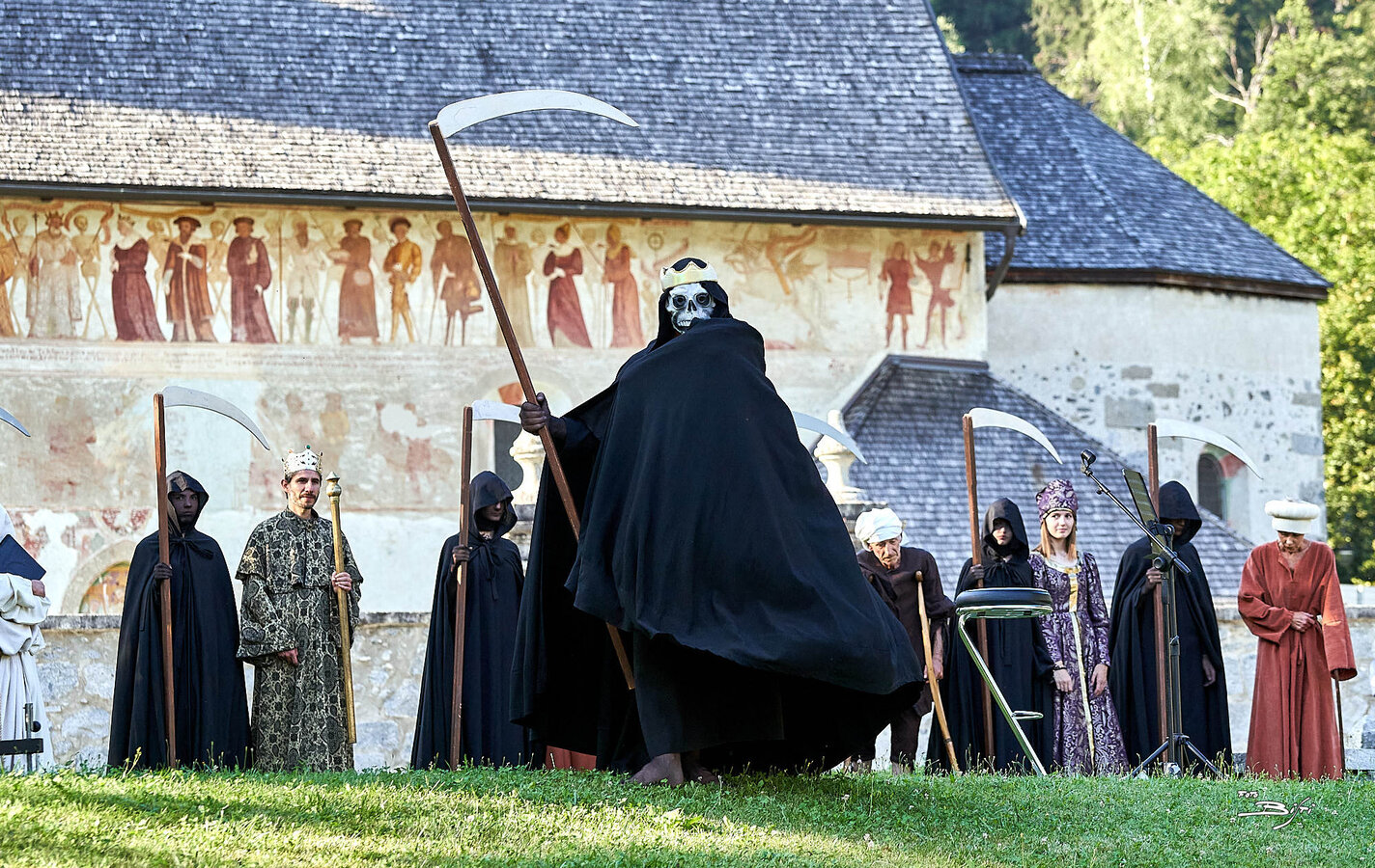 Theateraufführung mit vermummten Figuren und Sensen in einem historischen Innenhof.