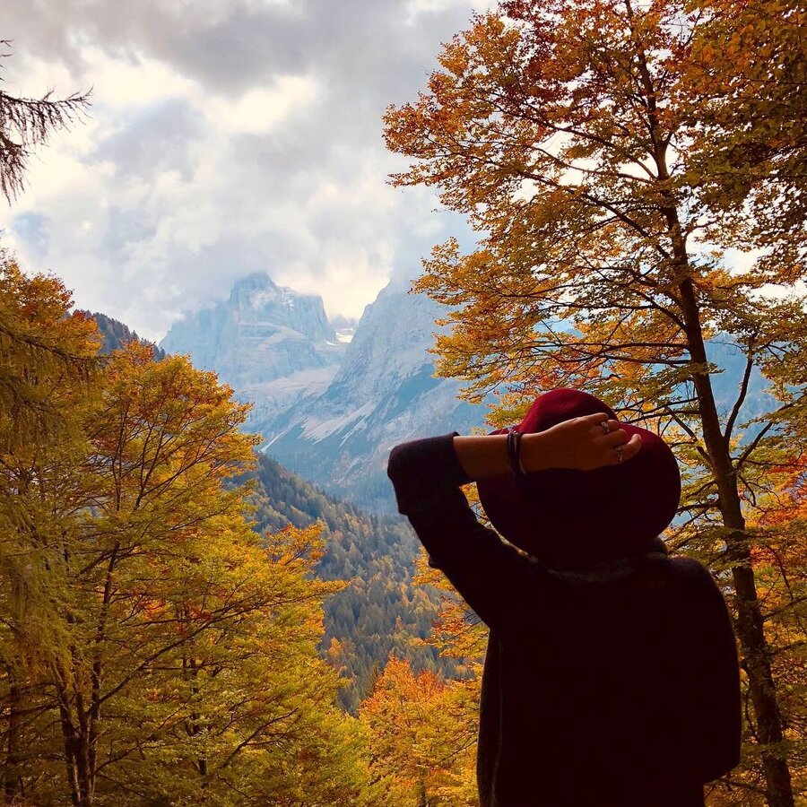 Persona con cappello rosso osserva montagne tra alberi autunnali colorati.