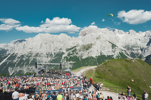 Folla a concerto all'aperto tra le montagne con parapendio nel cielo.